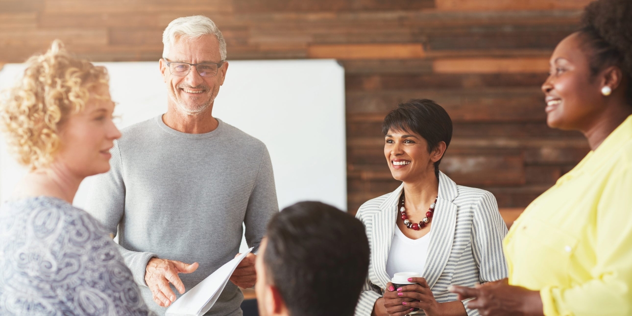 One white haired man wearing a light grey jumper talks to three woman (blond white woman in grey top, Asian woman with dark hair in grey jacket and black woman in a yello top) in an office setting with the back of another mans head visible at the bot